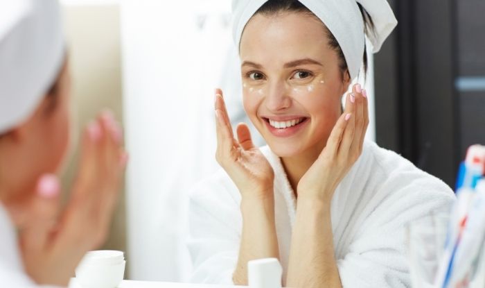 Mujer joven mirándose al espejo y sonriendo con manos en el rostro poniendo serum facial.